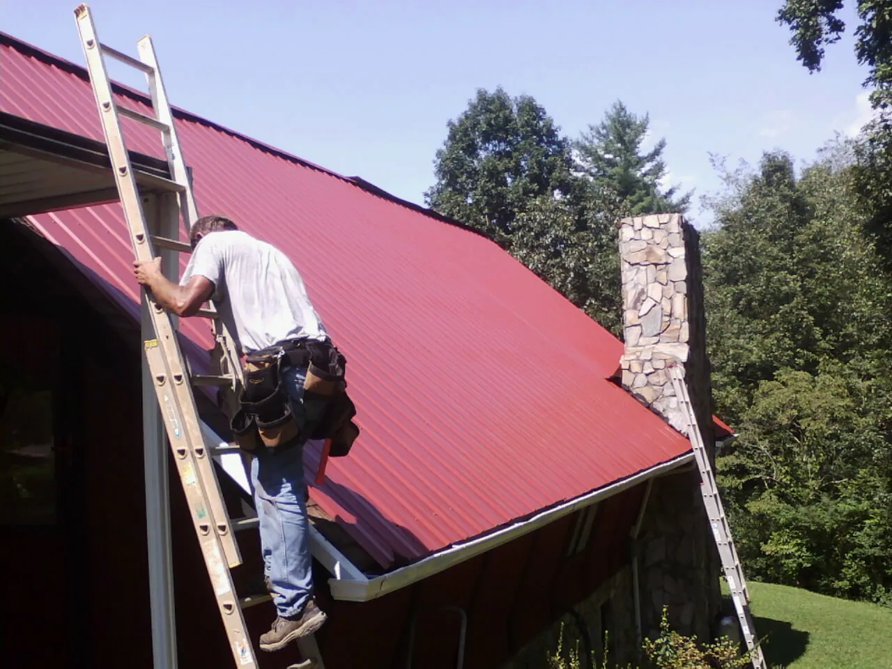 Roofer on a ladder beside a red metal roof and stone chimney — Soffit & Fascia Repair by Roofing Brothers in Cartersville